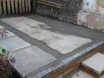 A freshly laid concrete shed base in a UK garden, showing an uneven grey surface with visible dips and patchy areas, bordered by timber edging bedded on fresh concrete