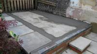 A freshly laid concrete shed base in a UK garden, showing an uneven grey surface with visible dips and patchy areas, bordered by timber edging bedded on fresh concrete