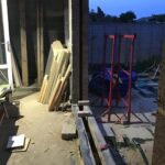 Rear house extension under construction at dusk, showing timber boards leaning against a blockwork wall, red acrow prop scaffolding in the garden, and an open cavity wall threshold with no door frame fitted