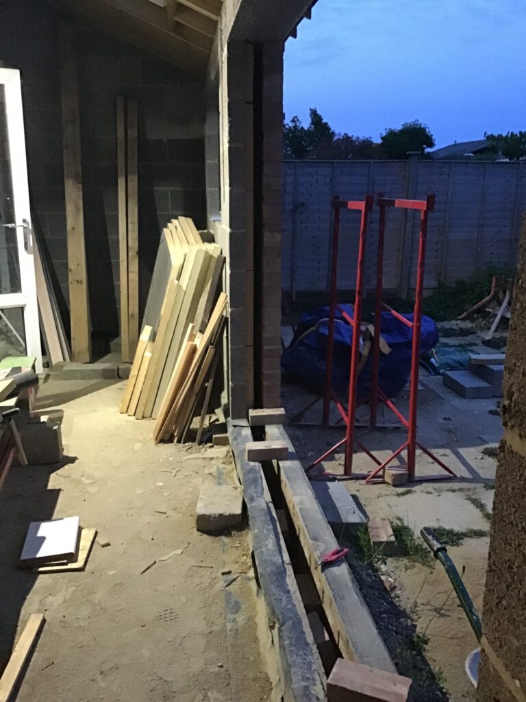 Rear house extension under construction at dusk, showing timber boards leaning against a blockwork wall, red acrow prop scaffolding in the garden, and an open cavity wall threshold with no door frame fitted