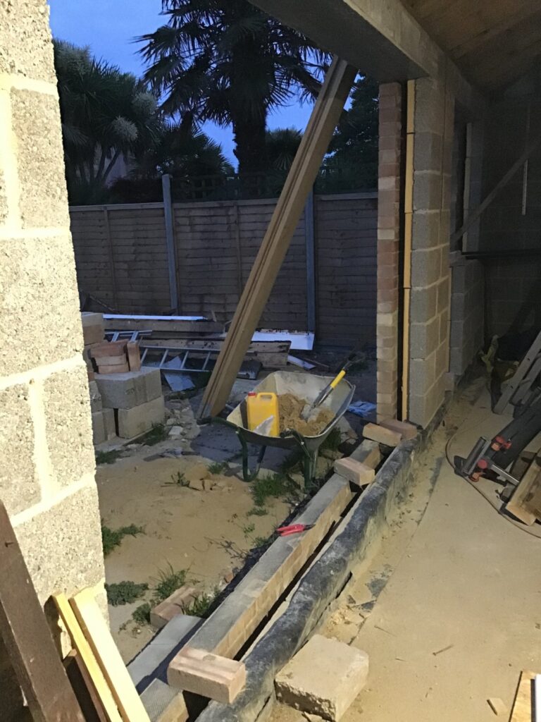Construction site of a rear extension at dusk, showing a cavity wall opening with brick and blockwork reveals, a timber prop bracing a board diagonally, and a wheelbarrow containing sand in the foreground