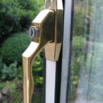 Gold-coloured brass uPVC window handle with keyhole lock fitted to a white double-glazed window frame, garden visible through the glass behind