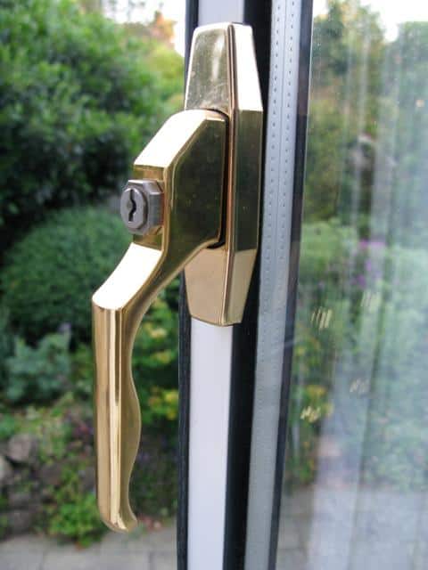 Gold-coloured brass uPVC window handle with keyhole lock fitted to a white double-glazed window frame, garden visible through the glass behind