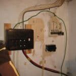 Old wooden-backed consumer unit with rewireable fuses mounted on timber batten, with green earth wire, red and black cables, and a separate fuse cutout on a bare plaster wall