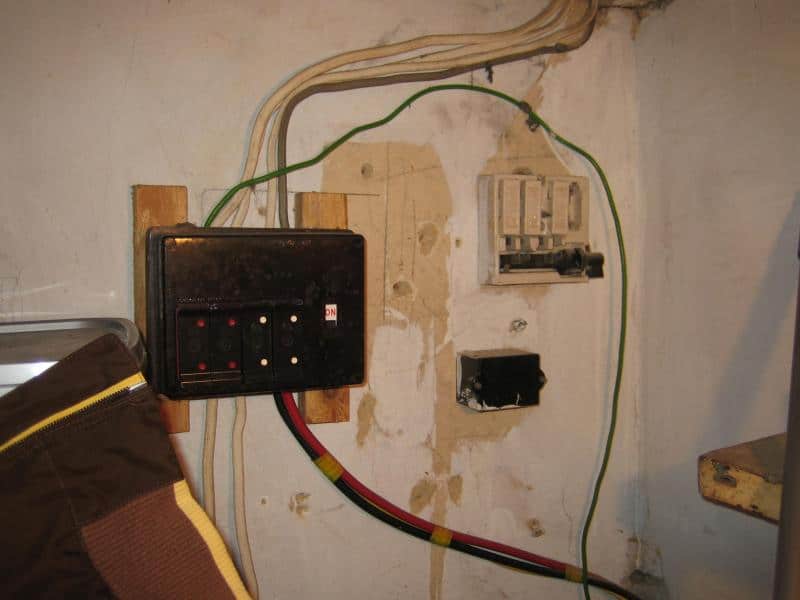 Old wooden-backed consumer unit with rewireable fuses mounted on timber batten, with green earth wire, red and black cables, and a separate fuse cutout on a bare plaster wall