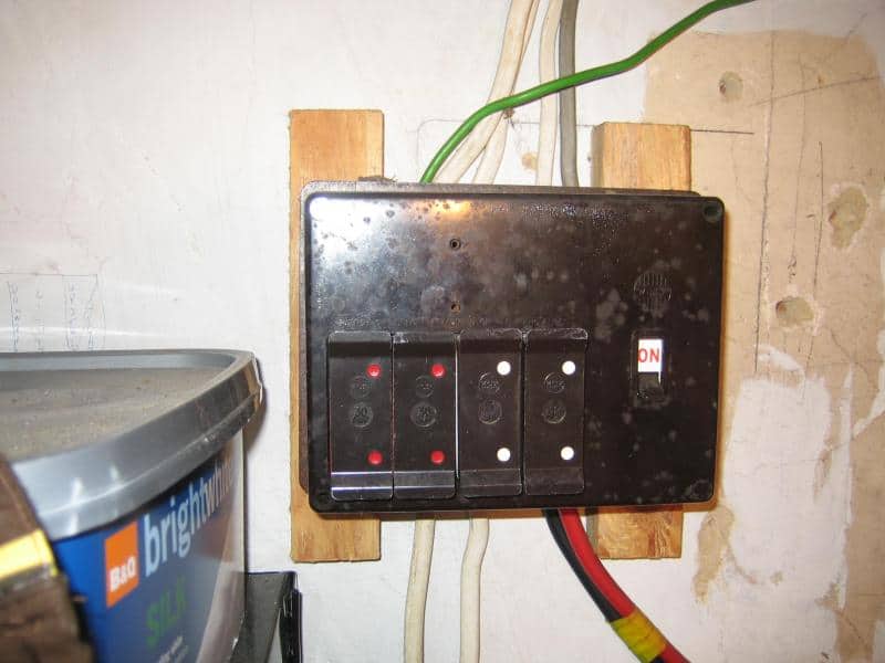 Close-up of a black plastic rewireable fuse board with four fuse carriers and an ON toggle switch, mounted on a timber batten with green, red, and black cables visible below