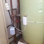 Green fibreglass hot water cylinder with copper pipework, a Grundfos circulating pump, and two white control units mounted on the wall beside it