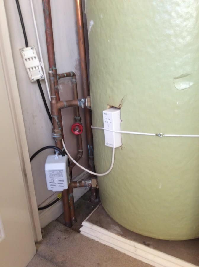 Green fibreglass hot water cylinder with copper pipework, a Grundfos circulating pump, and two white control units mounted on the wall beside it