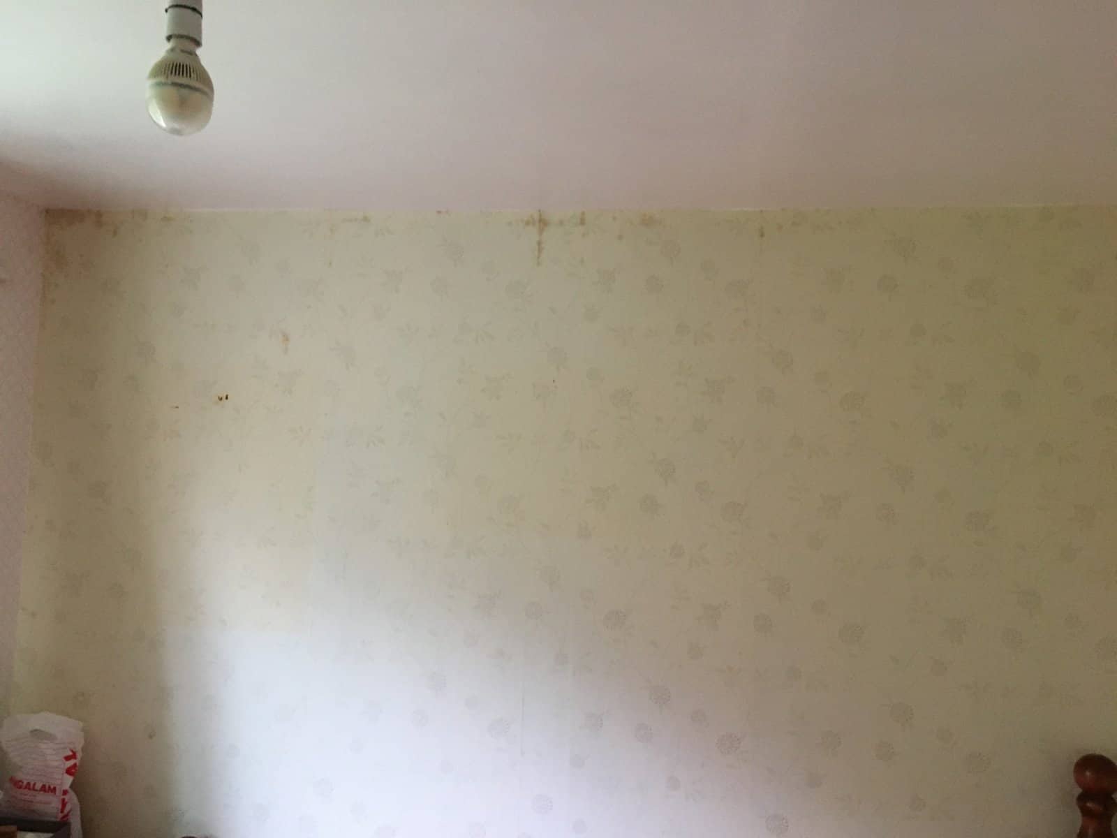 Interior view of an upstairs bedroom showing cream floral wallpaper with brown staining running in a band along the top of the external wall, fading towards the lower sections.