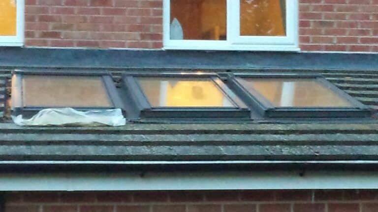 Three roof windows installed side by side on a low-pitch tiled roof, with a plastic sheet covering part of the left-hand window apron area