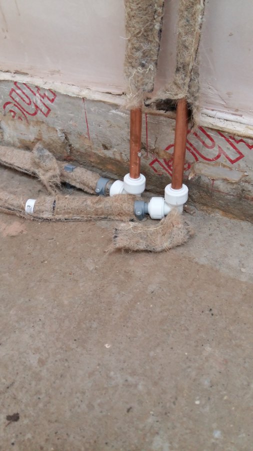 Two copper radiator tails rising from a concrete sub-base connected to grey flexible plastic pipes via white push-fit elbow fittings, with fibrous pipe lagging lying loose around the joints