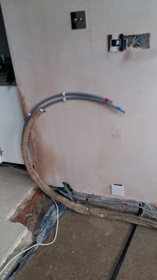 Grey flexible plastic pipes with push-fit fittings arching along a freshly plastered wall, with exposed electrical sockets and a partially lifted solid floor showing the sub-base beneath