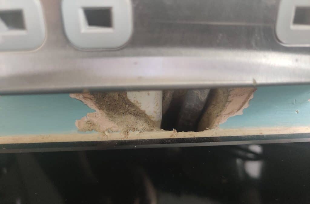 Close-up of a small chase cut into a teal kitchen wall just above a glass splashback, below an electrical socket, showing exposed plaster, dust and cables visible through the gap