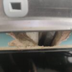 Close-up of a small chase cut into a teal kitchen wall just above a glass splashback, below an electrical socket, showing exposed plaster, dust and cables visible through the gap