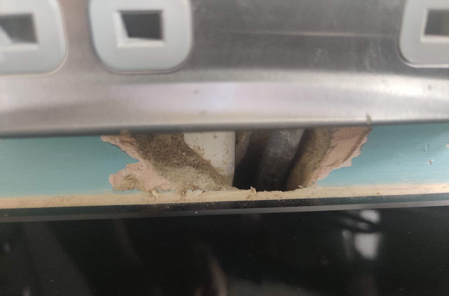 Close-up of a small chase cut into a teal kitchen wall just above a glass splashback, below an electrical socket, showing exposed plaster, dust and cables visible through the gap