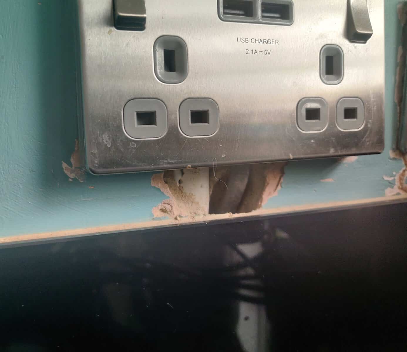 A double UK mains socket with USB charger ports mounted on a teal kitchen wall, with a ragged hole in the plaster visible beneath the socket faceplate just above the glass splashback