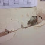 Peeling and crumbling lath and plaster wall showing multiple layers of paint and plaster separating from the substrate, with a calendar pinned above