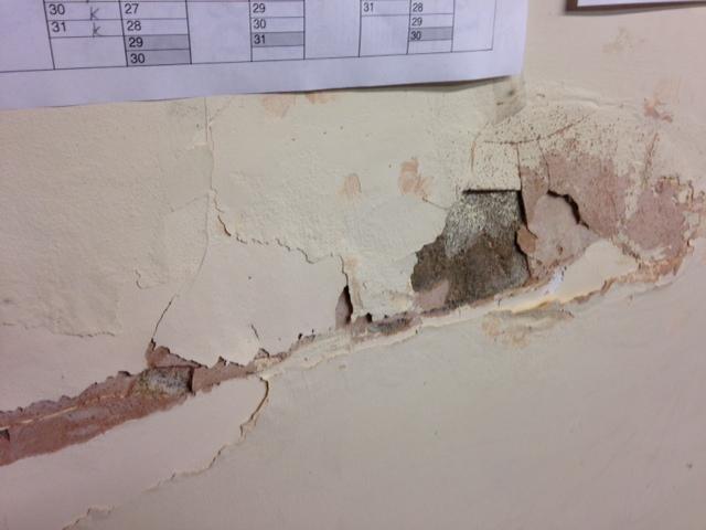 Peeling and crumbling lath and plaster wall showing multiple layers of paint and plaster separating from the substrate, with a calendar pinned above