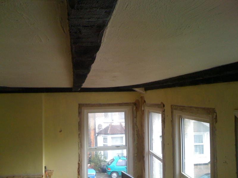 Lath and plaster ceiling partially collapsed, exposing the dark timber laths and structural joists above, in a room with bay windows