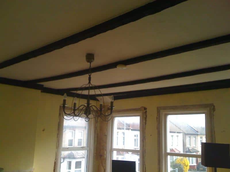 Replastered ceiling with dark timber beams in a period living room with bay windows and a wrought iron chandelier