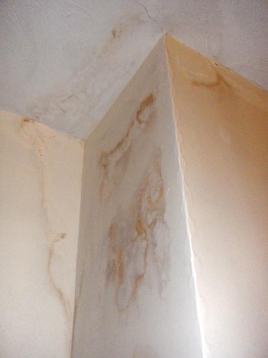 Brown water staining and damp patches spreading across a white painted chimney breast corner and ceiling in an upstairs room