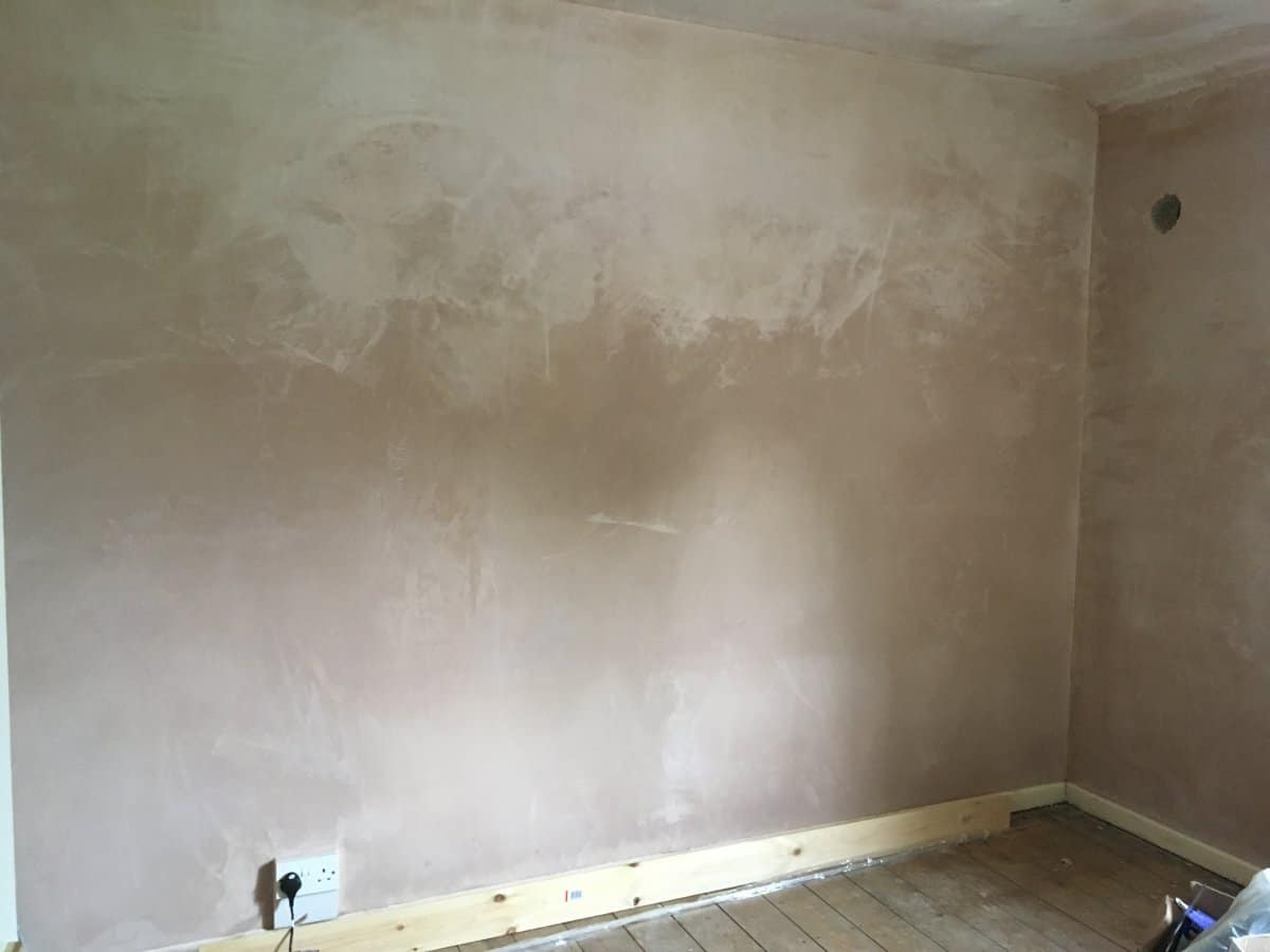 Freshly skimmed bedroom wall with uneven drying, showing pale dried patches alongside darker damp-looking areas, with bare wooden floorboards and a plug socket visible at the base.