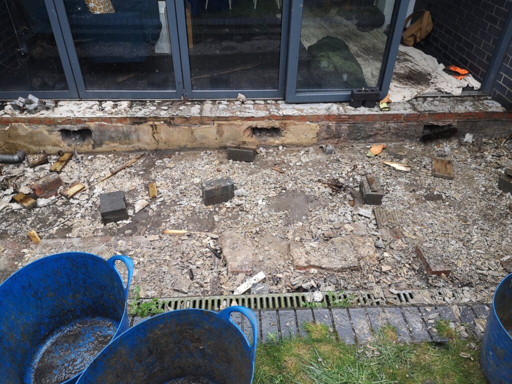 A demolished patio area outside sliding glass doors, showing rubble, broken concrete, scattered bricks, and a linear drainage channel, with blue builders' buckets in the foreground.