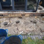 A demolished patio area outside sliding glass doors, showing rubble, broken concrete, scattered bricks, and a linear drainage channel, with blue builders' buckets in the foreground.