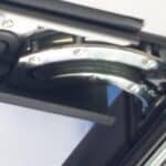 Close-up of a centre-pivot roof window friction hinge mechanism showing the curved metal pivot arm and fixing screws on a dark frame