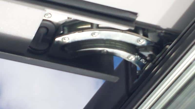 Close-up of a centre-pivot roof window friction hinge mechanism showing the curved metal pivot arm and fixing screws on a dark frame