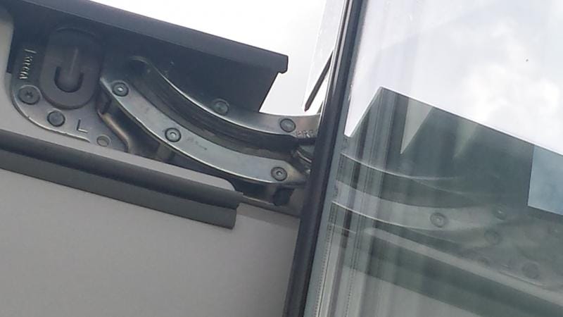 Centre-pivot roof window hinge assembly viewed from outside, showing the curved metal pivot bracket, adjustment screws, and glazing edge