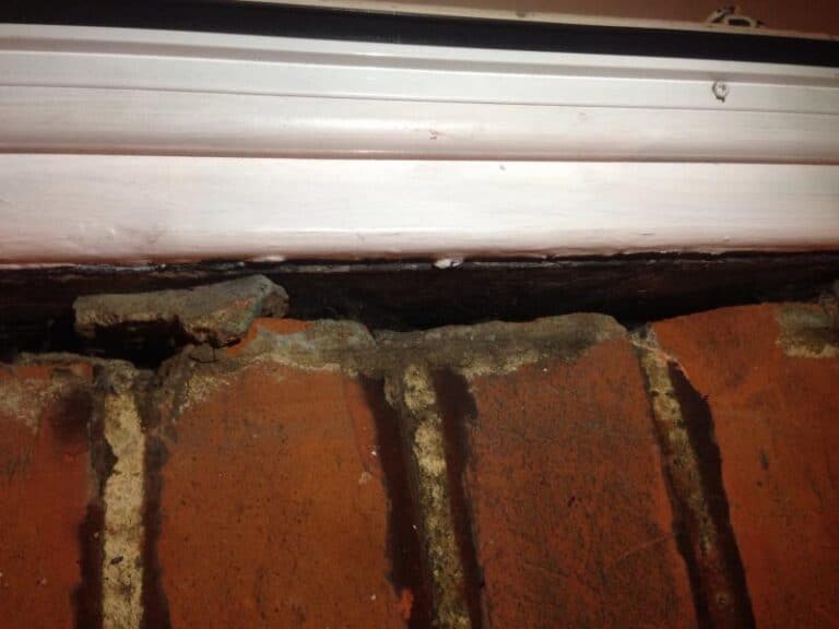 Close-up of a gap between a white uPVC door frame and the surrounding brickwork, showing crumbled mortar and exposed brick beneath the frame sill