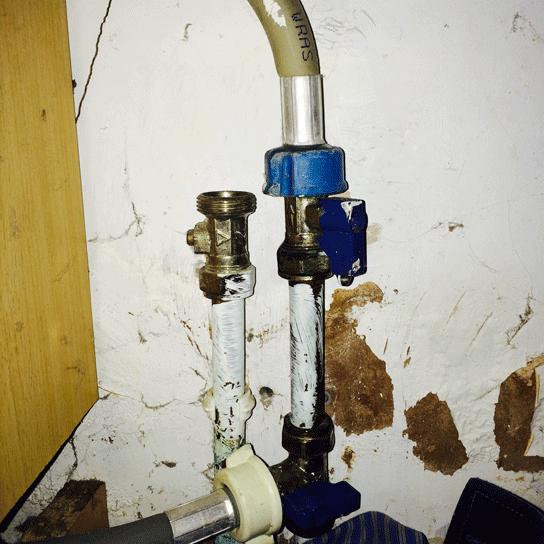 Close-up of a washing machine isolator valve with a blue quarter-turn lever handle, connected to a white braided hose and wrapped pipe fittings, mounted against a damp-stained wall.