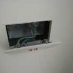 An open metal back box recessed into a plasterboard wall, showing blue, brown and earth wires connected to terminals, with the white twin socket face plate hanging loose below the box revealing a large gap between the box and the plasterboard surface