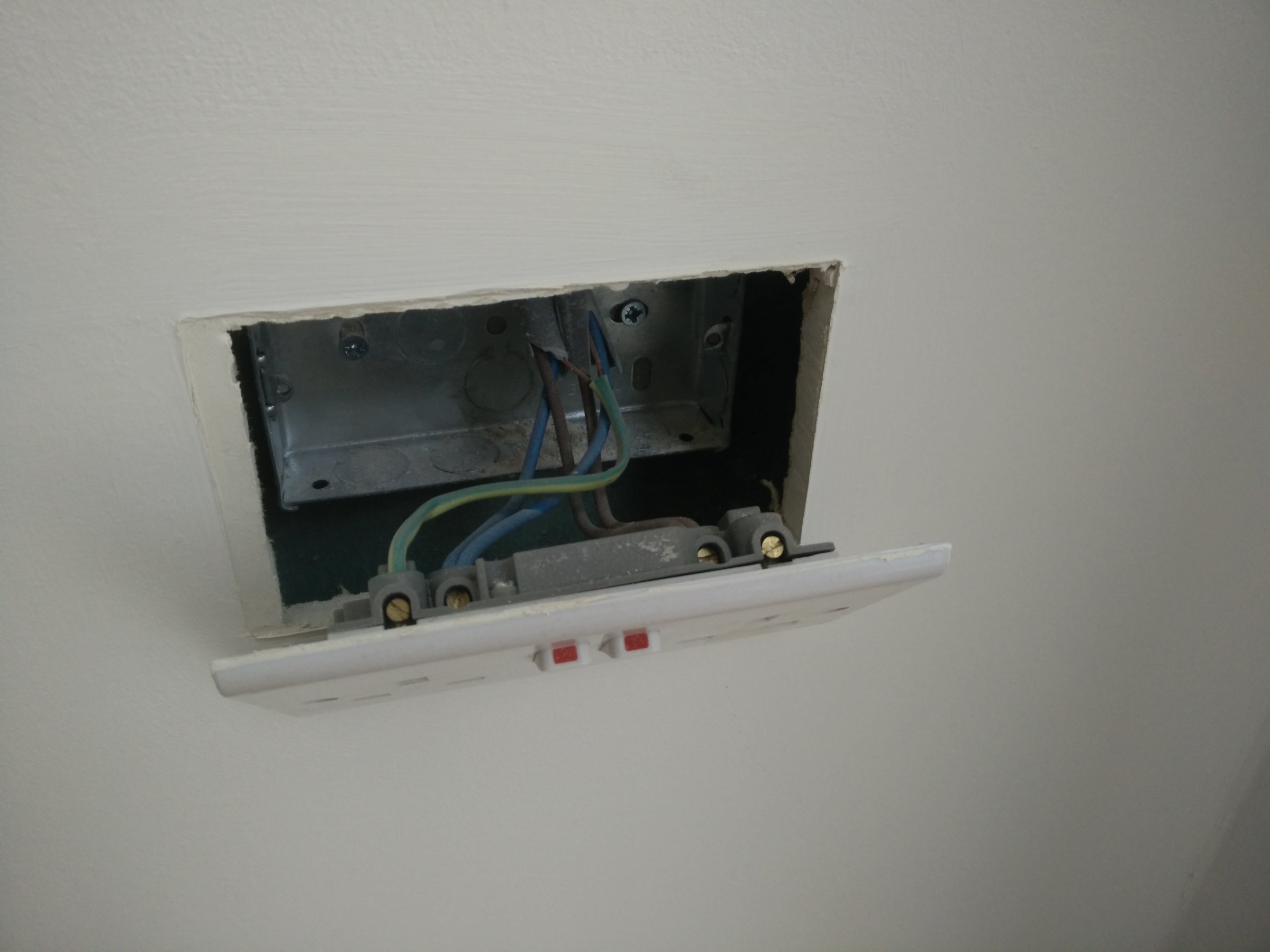 An open metal back box recessed into a plasterboard wall, showing blue, brown and earth wires connected to terminals, with the white twin socket face plate hanging loose below the box revealing a large gap between the box and the plasterboard surface