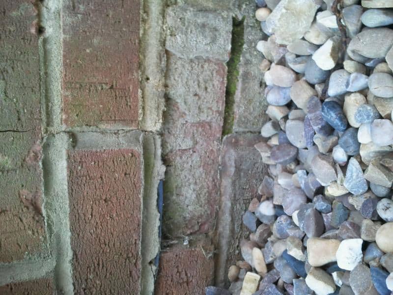 Close-up of a weathered red brick wall at ground level showing cracked mortar joints and a gap between the brickwork and a bed of gravel
