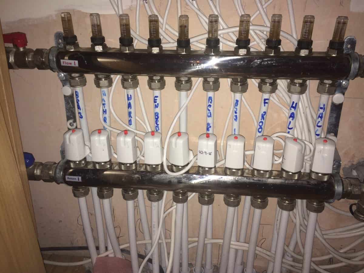 Close-up of UFH manifold flow and return bars with flow meters visible at the top, white actuators on the return ports, and pipes labelled in blue marker pen.