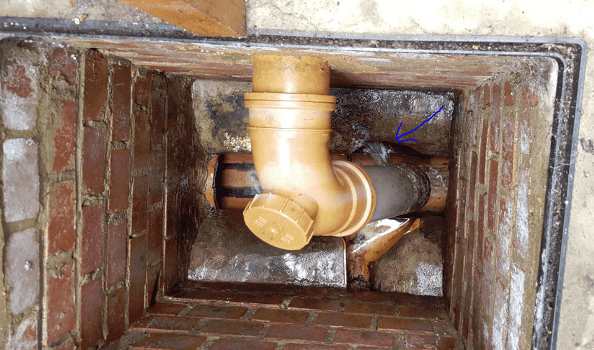 Inside a brick inspection chamber showing a new yellow uPVC swept bend with a rodding eye connected to an older clay drain pipe, with a blue arrow pointing to the junction.