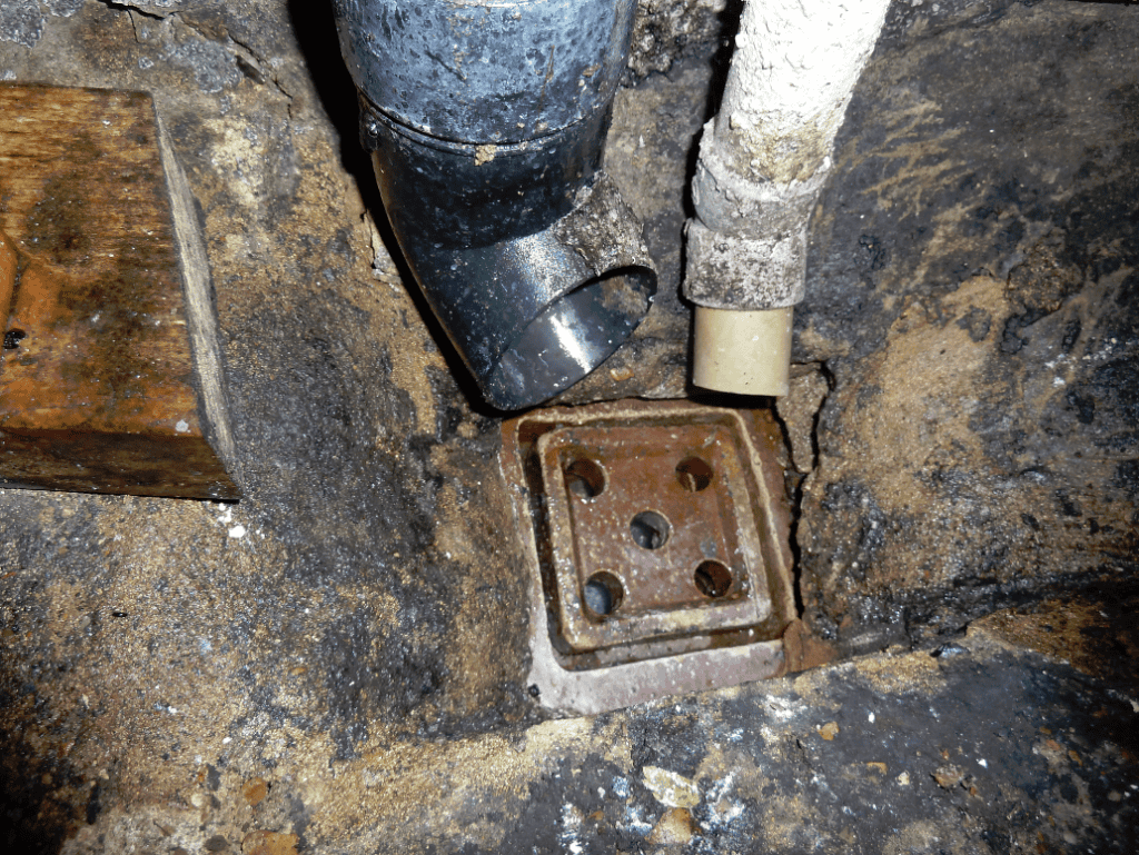 Old square cast-iron floor gully set into cracked concrete in a Victorian side return, with a black plastic soil pipe and a grey waste pipe discharging into it from above.