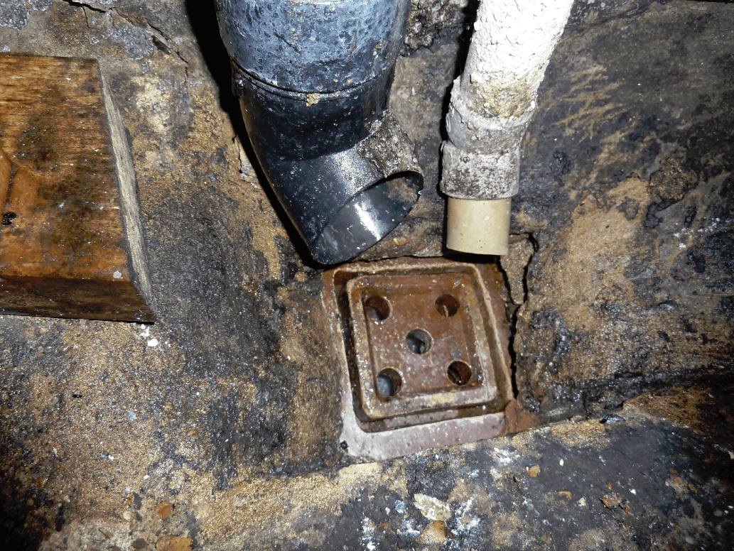 Old square cast-iron floor gully set into cracked concrete in a Victorian side return, with a black plastic soil pipe and a grey waste pipe discharging into it from above.