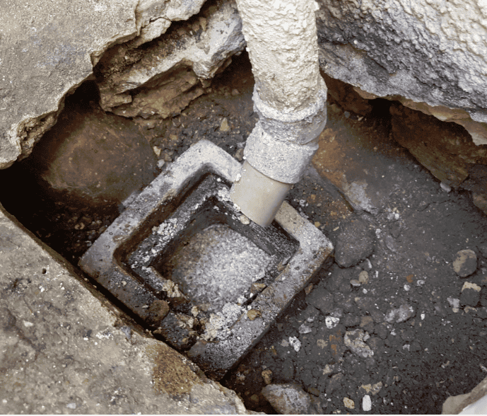 Close-up of a corroded square cast-iron gully trap with a grey plastic waste pipe entering it, surrounded by broken concrete and rubble.