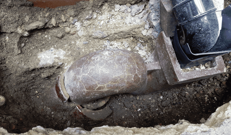 Excavated trench showing a badly cracked and cratered old clay drain pipe bend alongside a new black plastic push-fit fitting, ready for replacement.
