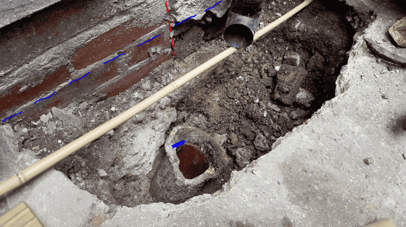 Excavated trench beside a brick wall showing a broken clay pipe at the base, a bamboo cane used as a probe, a black plastic downpipe above, and blue and red lines drawn on the image indicating the wall face and pipe alignment.