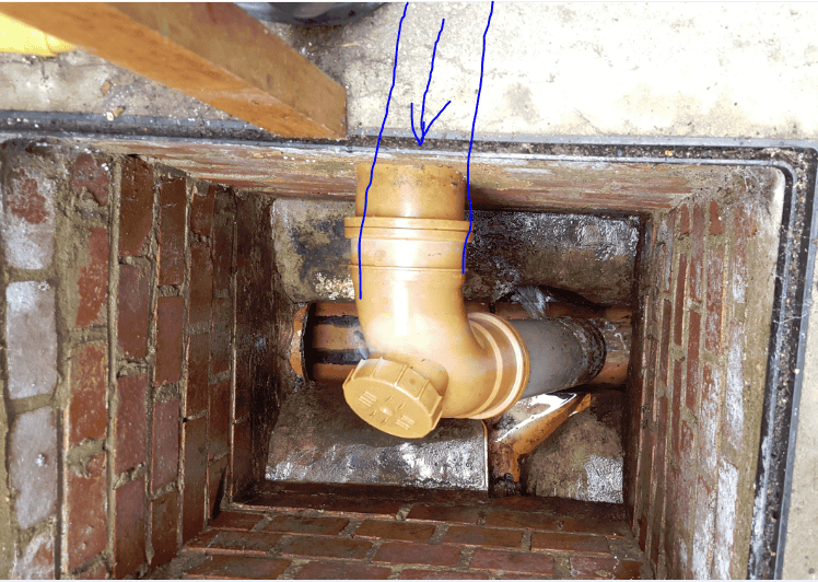 Inside a brick inspection chamber showing a new yellow uPVC 90-degree bend with a rodding eye, with blue arrows pointing to the pipe entry point at the top of the chamber.